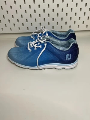 Blå sportskor från FootJoy - Snygga blå sportskor från FootJoy med vit sula och vita skosnören. Ovandelen är i meshmaterial för bra andningsförmåga och komfort. Skorna har en sportig design med FJ-logga på sidan och passar perfekt till träning eller golf.