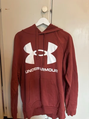hoodie från Under Armour - Snygg rostbrun hoodie från Under Armour med stor vit logga och text på bröstet. Hoodien har huva med dragsko, magficka och är gjord i mjukt ColdGear-material som håller dig varm. Perfekt för chill dagar eller träning.
