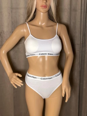 Vitt underklädesset från Calvin Klein M-L - Snyggt vitt underklädesset från Calvin Klein med sportig topp och matchande trosor. Båda delarna har elastiska band med logotypen tryckt runtom. Setet är gjort i mjuk bomull med stretch för en bekväm känsla. Perfekt för dig som gillar en enkel och stilren look.