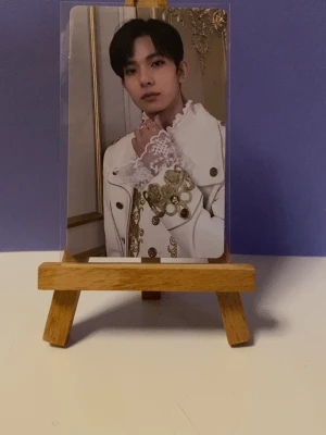 Heeseung border carnival photocard - Unikt fotokort med porträtt på framsidan och en grafisk, geometrisk baksida i svart, vitt och guld. Kortet är inramat i plastficka och står på ett litet staffli av trä. Perfekt för samlare eller som dekoration.