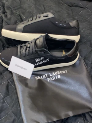 Saint Laurent svarta sneakers - Svarta Saint Laurent sneakers med vit sula och handskriven logga på sidan. Skorna har lågt skaft, svarta skosnören och detaljer i både läder och textil. Klassisk och stilren design med kontrasterande vit yttersula och diskreta detaljer. ⚠️Pris kan disskuteras vid snabbaffär⚠️