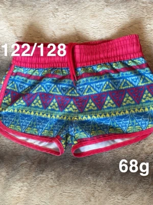 Färgglada mönstrade shorts med snörning - Säljer ett par färgglada shorts med blått, rött och gult mönster och röda detaljer. Shortsen har elastisk midja med snörning och är perfekta för sommaren. 🌸Fint använt skick. Storlek 122/128.