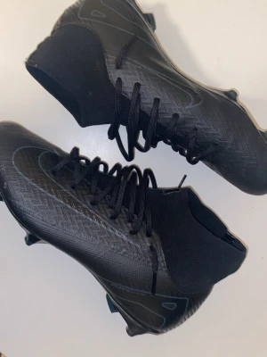 Svarta Nike Mercurial fotbollsskor - Säljer ett par helsvarta Nike Mercurial fotbollsskor med hög krage och snörning. Skorna har en modern, strömlinjeformad design och är tillverkade i syntetmaterial för bra grepp och kontroll på planen. Perfekta för dig som vill ha stilrena och snabba skor till matchen.