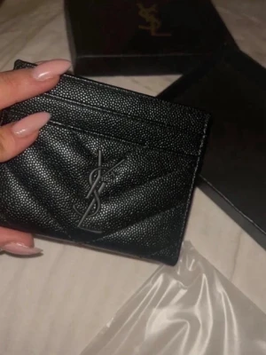 Svart YSL korthållare i läder - Säljer en svart korthållare från Saint Laurent med ikoniska YSL-loggan framtill. Tillverkad i strukturerat läder med quiltad design och flera kortfack. Perfekt för dig som vill ha en stilren och lyxig accessoar i fickan.