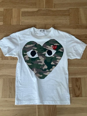 Vit Comme des Garçons PLAY t-shirt - Säljer en Comme des Garçons PLAY t-shirt i storlek S. Köpt på Selfridges i London. Vit t-shirt med camouflage-hjärtat framtill. T-shirten är i mycket bra skick och säljs eftersom den inte används längre. Hör av dig vid intresse eller frågor.