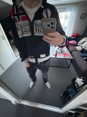 Svart hoodie från Geographical Norway - Säljer en svart hoodie från Geographical Norway med dragkedja, huva och stora tryck fram och bak. Flera broderade detaljer och patchar i vitt, rött och grått. Mjuk insida och ribbade muddar. Perfekt för dig som gillar streetwear och sportig stil.