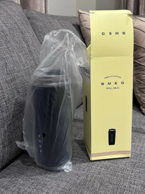 Svart Smeg Wall Mug med lock - Snygg svart Wall Mug från Smeg med stilren design och genomskinligt lock. Muggen är tillverkad i rostfritt stål och har Smeg-loggan tryckt vertikalt. Perfekt för att ta med kaffe eller te på språng. Levereras i originalförpackning.