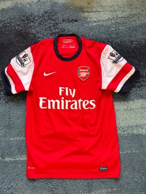 Arsenal matchtröja Nike röd/vit - Säljer en klassisk Arsenal matchtröja från Nike i rött och vitt med klubbmärke och Fly Emirates-tryck på bröstet. Tröjan har svarta detaljer vid krage och ärmslut samt Premier League-patch på ärmarna. Tillverkad i Dri-FIT material som andas. Är äkta köpt från deras stadium 