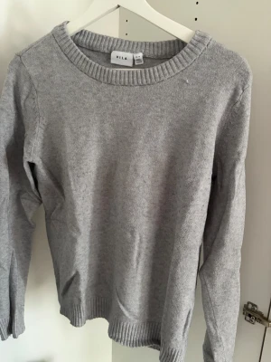 Grå stickad tröja från VILA - En enkel och stilren grå stickad tröja från VILA. Tröjan har rund halsringning, långa ärmar och ribbade muddar vid ärmslut och nederkant. Perfekt att matcha med jeans eller kjol för en avslappnad look.