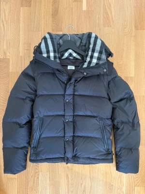 Svart pufferjacka från Burberry - Snygg svart pufferjacka från Burberry med avtagbar huva och klassiskt rutigt foder. Jackan har dragkedja och tryckknappar framtill, två snedställda fickor med dragkedja och en boxig, puffig passform. Armarna går att ta bort då jackan blir en väst. Perfekt för kalla dagar och riktigt stilren. Kvitto finns!