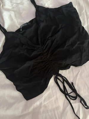 Svart croppad linnetopp med snörning - Snygg svart croppad linnetopp från Gina Tricot med tunna axelband och trendig snörning framtill som kan justeras. Toppen har en lätt rynkad effekt i mitten och är perfekt för sommarens alla outfits. Materialet känns mjukt och lätt mot huden.