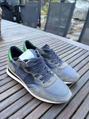 Gråblå sneakers från Philippe Model - Snygga sneakers i grått och blått från Philippe Model med detaljer i mocka och mesh. Skorna har gröna detaljer vid hälen, vita sulor och mörkblå skosnören. Klassisk låg modell med sportig vibe och diskret logga på sidan.
