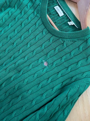 Grön kabelstickad tröja från GANT - Snygg grön kabelstickad tröja från GANT med rund halsringning och diskret broderad logga på bröstet. Tröjan har ribbade muddar och är tillverkad i mjukt material som känns skönt mot huden. Perfekt för dig som gillar klassisk och stilren look.