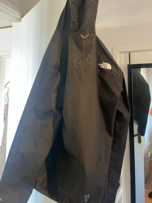 Svart vindjacka från The North Face - Snygg svart vindjacka från The North Face med huva och diskret mönster på axelpartiet. Jackan har logga på bröstet och är tillverkad i ett lätt, vattenavvisande material. Perfekt för blåsiga dagar och enkel att packa ner i väskan. Pris kan diskuteras 