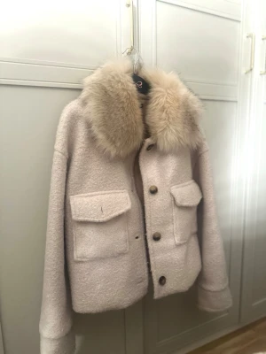 Beige pälsjacka med stor krage - Säljer en beige pälsjacka med stor fluffig krage i fuskpäls och två stora fickor framtill. Onesize