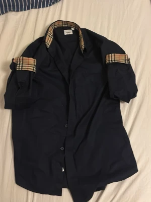 Mörkblå burberry skjorta  - Säljer en mörkblå skjorta från Burberry med klassiska beige-rutiga detaljer på krage och ärmslut. Skjortan har knappar framtill och kan bäras med uppvikta ärmar för en avslappnad look. Knappen högst upp är sönder men annars är den i fint skick. Den är i storlek M men sitter som S