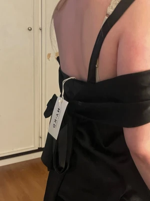 Svart omlott tubtopp från NA-KD - Säljer en svart tubtopp från NA-KD med omlott-detalj och knyt i ryggen. Toppen är axelbandslös och har en elegant, slät finish som passar perfekt till olika outfits. Materialet känns mjukt och har en lätt glans.