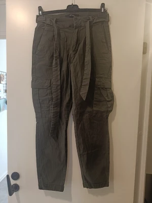 Mörkgråa cargobyxor med knytband - Säljer ett par mörkgråa cargobyxor med breda ben och stora fickor på sidorna. Byxorna har ett matchande knytband i midjan och är tillverkade i ett mjukt, lite skrynkligt tyg som ger en avslappnad look. Perfekta för en chill och trendig stil.