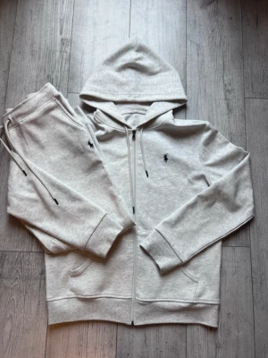 Grått loungewear-set från Polo Ralph Lauren - Mysigt loungewear-set i ljusgrått från Polo Ralph Lauren. Setet består av en hoodie med dragkedja och fickor samt matchande byxor med dragsko i midjan. Båda plaggen har den klassiska mörkblå Polo-loggan broderad på bröstet och benet. Perfekt för chill dagar hemma.