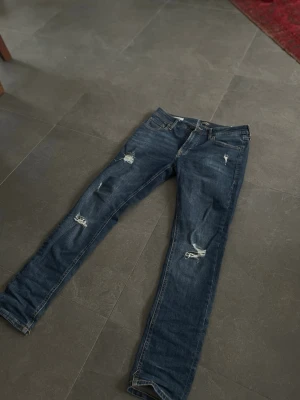 Blå slim jeans med slitningar  - Säljer ett par blå slim jeans med slitningar och hål på benen för en trendig och avslappnad look. Jeansen har klassisk femficksdesign och knappgylf. Perfekta för dig som gillar en streetstyle vibe. Väldigt clean jeans allmänt och passar till allt😃 priset är inte hugget i sten!!