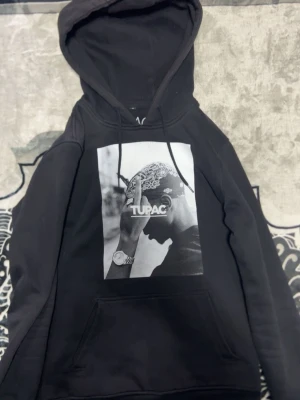 Svart Tupac hoodie Mister Tee x ARTISTAR - Svart hoodie med stor svartvit Tupac-print på bröstet och texten 'TUPAC'. Klassisk känguruficka och huva med dragsko. Tillverkad i mjukt bomullsmaterial, perfekt för en streetwear-look.