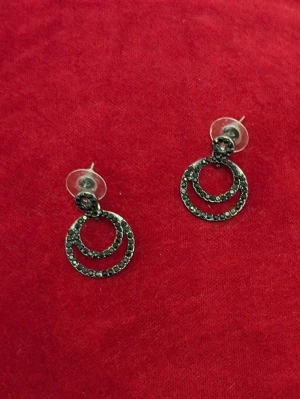 Svarta cirkelformade örhängen - Säljer ett par snygga örhängen i svart metall med dubbla cirklar och små gnistrande stenar. Örhängena har stift och transparenta bakstycken. Perfekta för dig som gillar en modern och edgy stil.