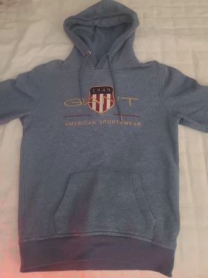 Blå hoodie från Gant med tryck - Snygg blå hoodie från Gant med broderad logga och texten 'American Sportswear' på bröstet. Klassisk känguruficka framtill, dragsko i huvan och ribbade muddar. Perfekt för en avslappnad och sportig stil.