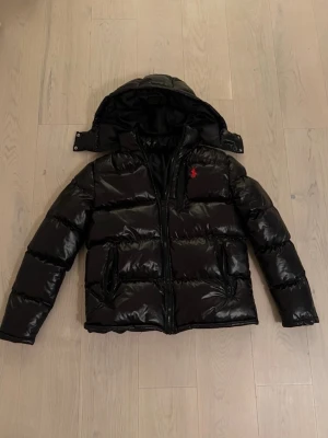 Svart pufferjacka från Ralph Lauren - Snygg svart pufferjacka från Ralph Lauren med blank finish och avtagbar huva. Jackan har två dragkedjeförsedda fickor och röd broderad logga på bröstet. Ej autentiskt, därav priset. Inga tags inuti jackan utan endast en liten storlekslapp. Vid frågor är det bara att höra av sig✅✅ INGEN RETUR❌