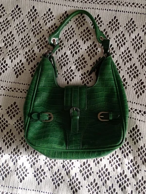 Grön handväska - Supersnygg grön krokodilmönstrad väska med silvriga detaljer. Magnetstängning och dragkedja. Inuti finns ett dragkjedjefack. Även dragkjedja på baksidan med en söt stjärna. Innerfodret är tyvärr trasigt men väskan är fullt användbar ändå och annars bra skick🩷