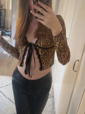 Leopardmönstrad croppad topp med snörning - Snygg croppad topp med leopardmönster och långa ärmar. Toppen har v-ringning, svarta spetsdetaljer och knyt framtill. Perfekt för dig som vill sticka ut med en trendig och djärv look.