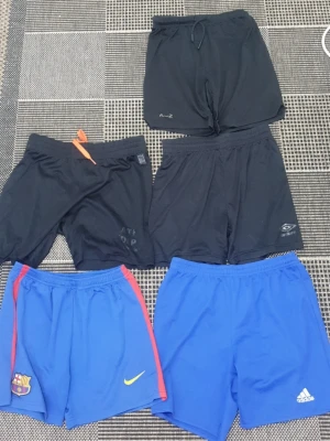 5-pack sportshorts Nike, Adidas, m.fl. - Säljer ett 5-pack sportshorts i svart och blått från Nike, Adidas, Barcelona, A-Z och Umbro. Perfekta för träning eller fotboll. Alla har elastisk midja, vissa med snörning. Materialet är lätt och andas, troligen polyester.