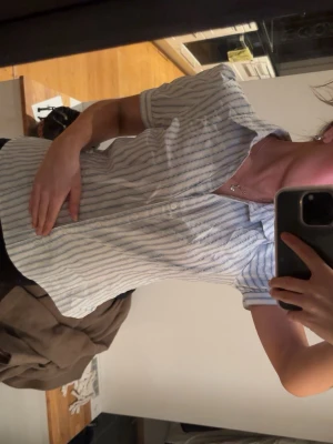 Vit kortärmad randig skjorta - Säljer en vit skjorta med tunna blå ränder och korta ärmar. Skjortan har klassisk krage och knäppning framtill. Perfekt för en clean och fräsch look. Materialet känns lätt och svalt, troligen bomull eller en bomullsblandning.