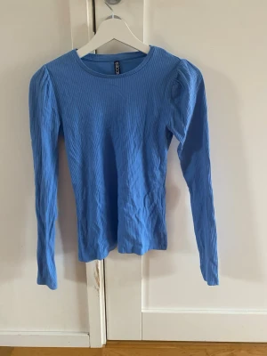 Blå ribbad långärmad topp Pieces - Säljer en ljusblå ribbad långärmad topp från Pieces i storlek XS. Toppen har puffade axlar och är tillverkad i mjuk bomullsmix. Perfekt att styla med jeans eller kjol för en trendig look.