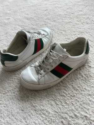 Gucci ace sneakers vita - Tja säljer ett par Gucci ace skor, vita i storlek 43. De vill säga typ 8 i Gucci storlek. Det är den gamla modellen riktigt snygga skor går till varje outfit. Snörerna har blivit lite missfärgade och de är lite crees men de är inget som inte går att fixa jätte änkelt på en sko tvätt. Brukar finnas inne i stan. Säljer för jag vill köpa ett par andra så om ni har några funderingar så hör av er. Sjävklart är dem äkta. Tyvärr så finns inte Box eller kvitto kvar men dom som har ett öga för Gucci ser