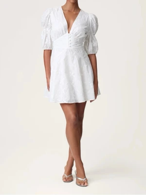 Vit kort klänning - Söt vit kortklänning med puffärmar och v-ringning. Klänningen har knappar framtill, markerad midja och ett diskret blommigt mönster i tyget. Aldrig använd med lappen kvar💗kan skicka fler bilder!