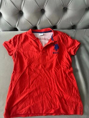 Röd pikétröja från US Polo Assn (pojke) - Snygg röd pikétröja från US Polo Assn med blå logga och text på bröstet. Klassisk krage med knappslå och korta ärmar. Perfekt till jeans eller shorts för en sportig look. Storlek 11-12 år eller 146-152 cm