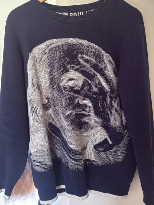 Mörkblå stickad tröja från Soul London - Mörkblå stickad tröja från Soul London med stor vit och grå konstnärlig ansiktsillustration på framsidan. På ryggen står texten 'MOTIVATED BY THE FEAR OF BEING AVERAGE' i vitt. Tröjan har rund hals och ribbade muddar i vitt och blått.