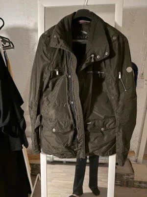 Mörkgrön vindjacka från Snoot - Stilren mörkgrön vindjacka från Snoot med flera fickor, dragkedjor och tryckknappar. Jackan har hög krage, lång ärm och detaljer i silverfärg. Perfekt för dig som gillar funktionella och snygga ytterplagg.