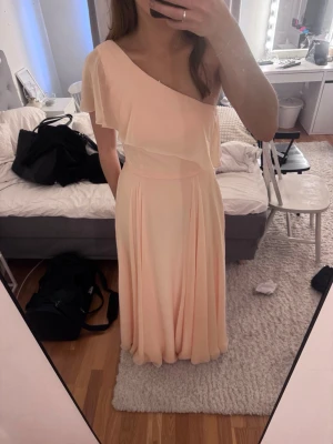 Ljusrosa oneshoulder långklänning - Elegant ljusrosa långklänning med oneshoulder och volangdetalj över axeln. Klänningen har ett svepande fall och markerad midja, perfekt som exempelvis balklänning. Köpt för 1500kr, säljer då jag hittat en annan inför balen 