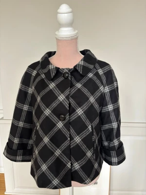 Svartvit rutig kappa med stora knappar - Stilren svart kappa med vitt rutmönster och stora dekorativa knappar framtill. Kappan har bred krage, trekvartsärmar med uppvik och en snygg detalj baktill med knappar. Storlek 42 men passar S/M