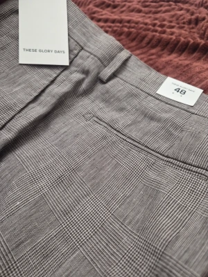 These Glory Days T Noble Pow Check ritiga byxor, storlek 48 - Snygga kostymbyxor från These Glory Days i grått med klassiskt rutmönster. Byxorna har raka ben, bälteshällor och diskreta bakfickor. Perfekta för en stilren och modern look. Materialet känns mjukt och följsamt.