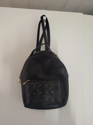 Svart ryggsäck med nitar - Snygg svart ryggsäck i konstläder med guldfärgade dragkedjor och små guldfärgade nitar på ytterfickan. Ryggsäcken har justerbara axelband och ett praktiskt handtag upptill. Perfekt för dig som vill ha en stilren men edgy look.