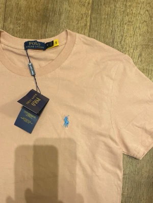 Rosa t-shirt från Polo Ralph Lauren (Helt ny / Oanvänd) - Säljer en rosa t-shirt från Ralph Lauren med klassisk blå logga broderad på bröstet. T-shirten har rund halsringning och korta ärmar. Tillverkad i mjuk bomull för en skön känsla och enkel stil. Storlek Medium men passar mer som small. Snabb Leverans!