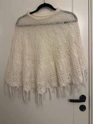 Vit stickad poncho från KappAhl - Mysig vit poncho sjal från KappAhl med vackert hålmönster och fransar längs kanten. Poncho har en rundad form och är perfekt att slänga över axlarna för extra värme. Tillverkad i mjukt garn som känns skönt mot huden.