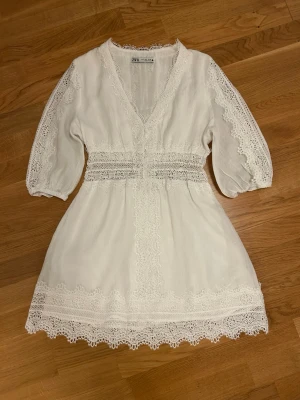  Ny vit  spetsklänning från Zara - Superfin vit kortklänning från Zara med v-ringning, spetsdetaljer och trekvartsärm. Klänningen har vackra broderade spetskanter längs ärmar, midja och nederkant samt omlottkänsla framtill. Perfekt för sommarens alla tillfällen!