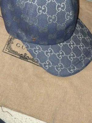 Blå GG-mönstrad keps från Gucci - Säljer en blå keps från Gucci med det klassiska GG-monogrammet över hela kepsen. Kepsen är i tygmaterial och har böjd skärm. Perfekt för dig som vill ha en exklusiv och trendig accessoar med tydlig logga.