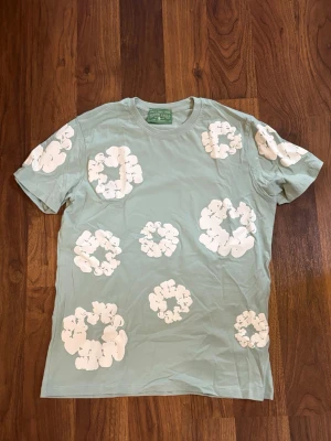 Grön t-shirt från denim tears - Snygg ljusgrön t-shirt från denimtears med stora vita blommiga tryck över hela tröjan. Klassisk rund hals och korta ärmar. Perfekt för dig som vill sticka ut med ett coolt mönster och mjukt bomullsmaterial. Skriv vid frågor
