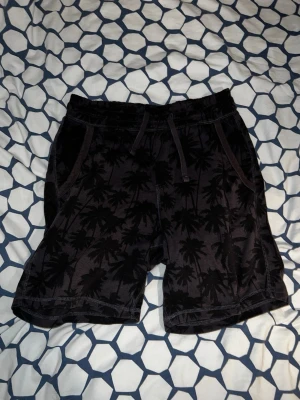 Svarta shorts med palmtryck och snörning - Säljer ett par svarta shorts med palmmönster och snörning i midjan. Shortsen har två sidofickor fram och en bakficka. Perfekta för sommaren och riktigt sköna att bära. Materialet känns mjukt och bekvämt.