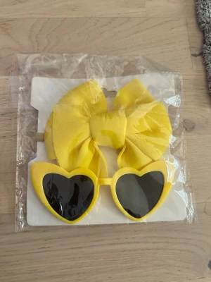 Gula hjärtformade solglasögon med pannband/hårband - Gula hjärtformade solglasögon med pannband/hårband Säljer ett par solglasögon med hjärtformade bågar i starkt gul färg och mörka linser. Perfekt accessoar för dig som vill sticka ut och addera lite färg till din stil. Medföljer även en stor gul rosett som matchar glasögonen.