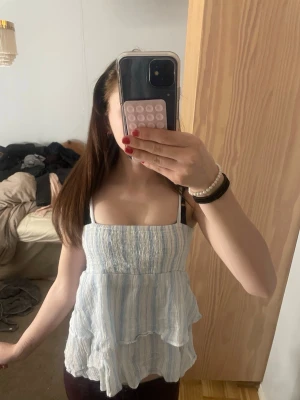 Ljusblå randig volangtop - Supersöt ljusblå och vit randig linnetopp med volangdetaljer och breda axelband. Toppen har en luftig och flowy passform, perfekt för varma dagar. Materialet känns mjukt och svalt, troligen bomull eller linne. Perfekt att styla med jeans eller kjol. Hör järna av dig för frågor💕❤️